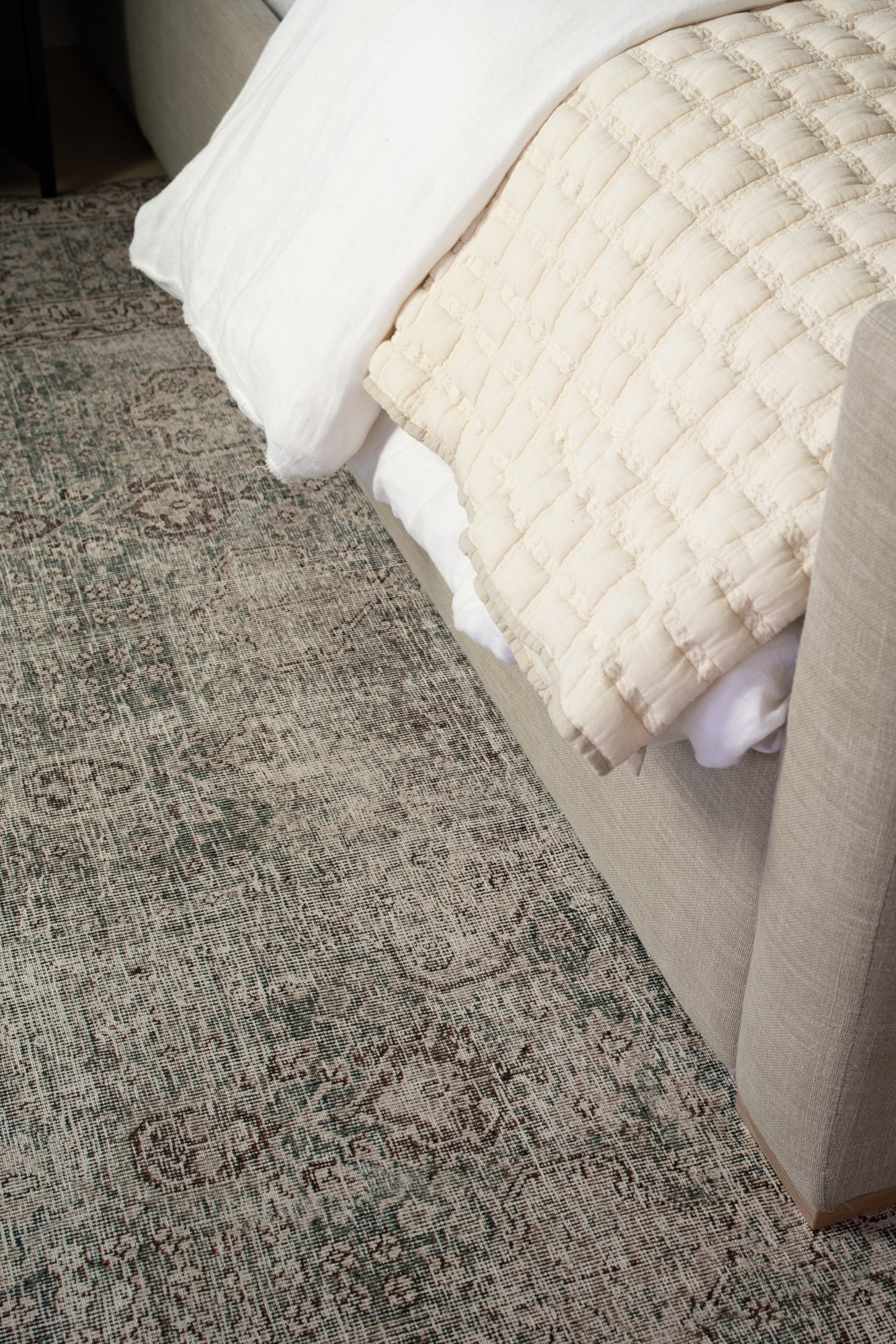 A cozy beige couch with a quilted cream blanket drapes over the arm. It sits beside an intricately patterned, muted-toned area rug on a wooden floor.