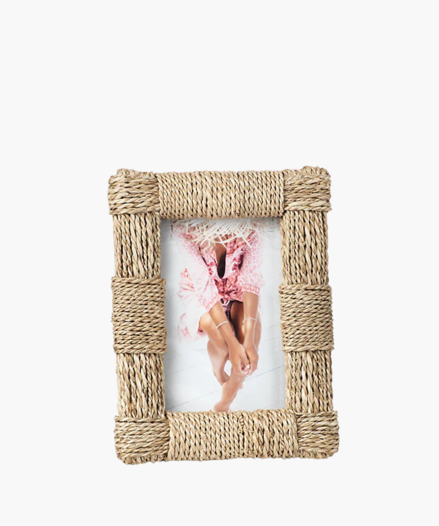 A rustic, woven rope frame with a picture of a person in a pink floral dress and straw hat sits on the floor, with legs bent and arms resting on knees.