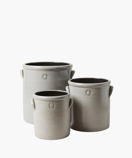 Three gray stoneware crocks of varying sizes with small handles. Each crock has a subtle embossed wreath design, conveying a rustic, artisanal feel.