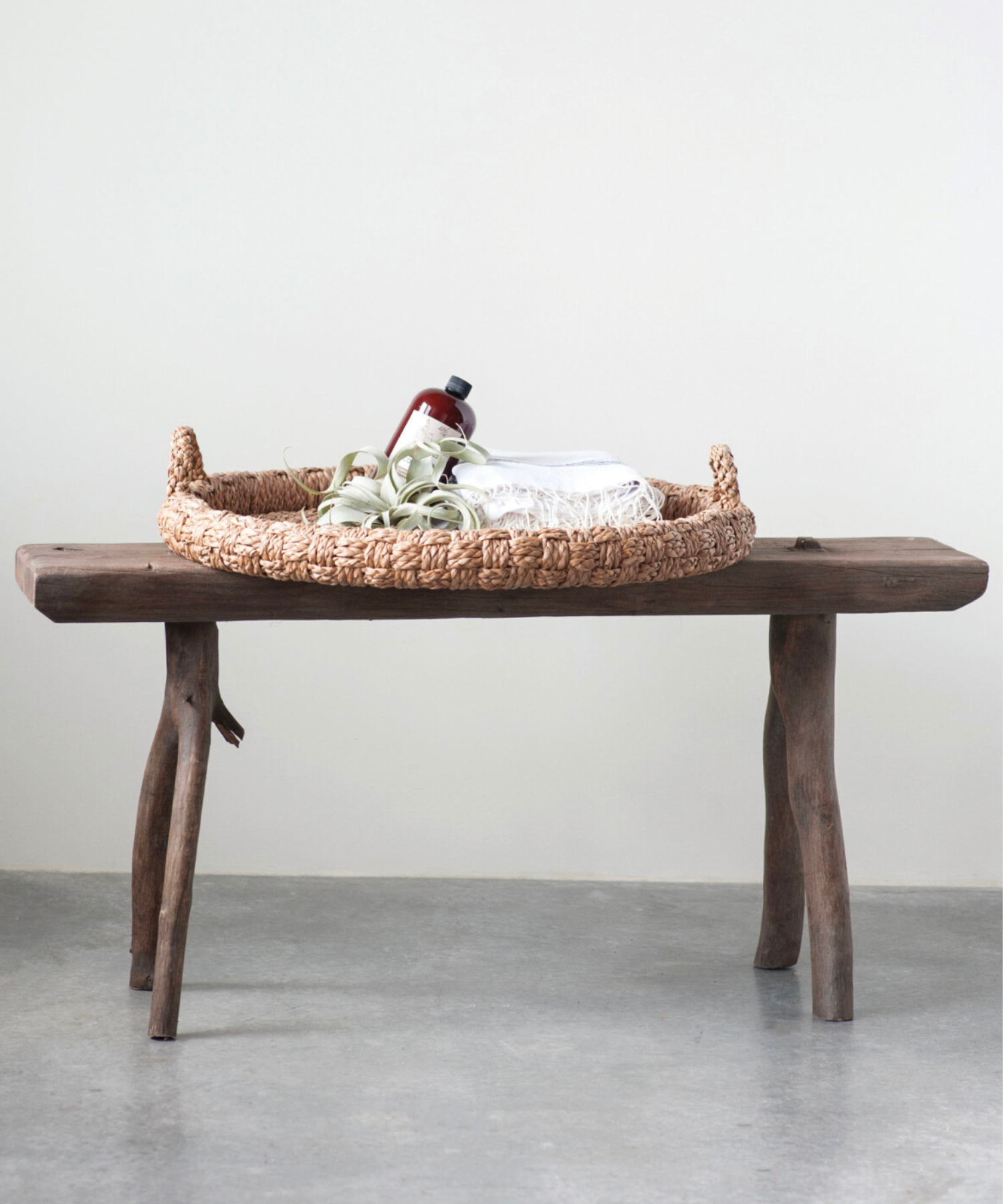 Round braided wicker tray or basket with two looped handles styled on a wooden bench. The natural fibers create a warm, rustic feel.