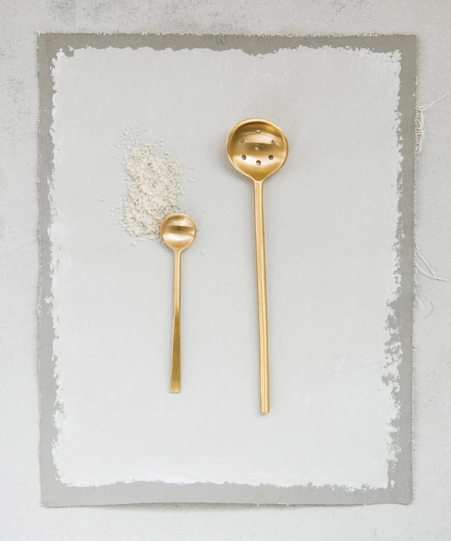 A minimalist brass slotted spoon with a long handle and round, perforated ladle styled with a small brass spoon, evoking elegance and simplicity.