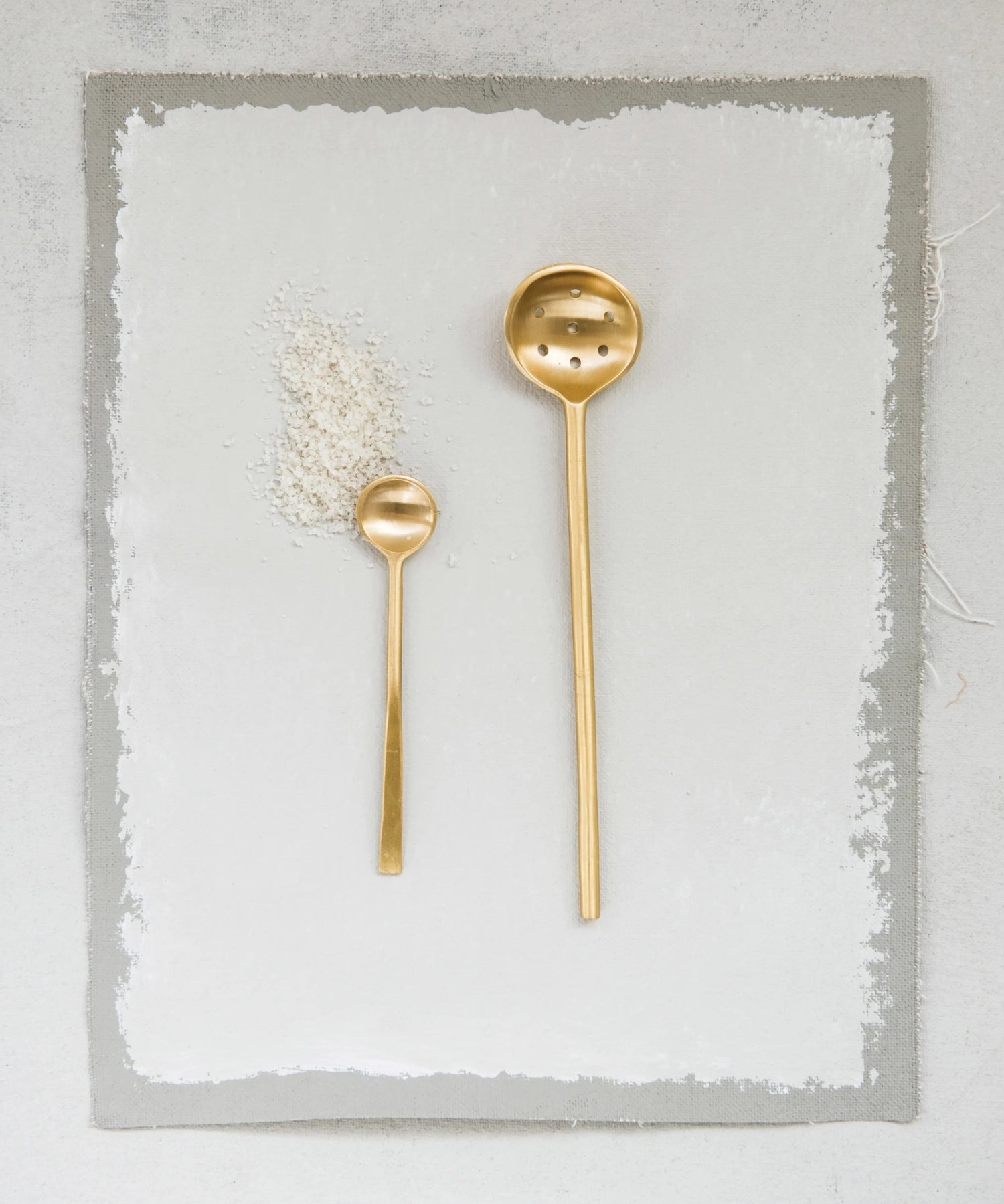 A minimalist brass slotted spoon with a long handle and round, perforated ladle styled with a small brass spoon, evoking elegance and simplicity.