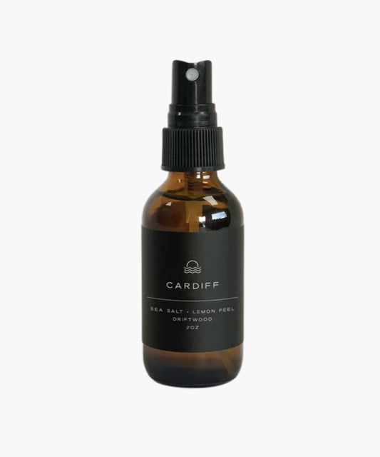 2oz amber spray bottle with black label reading "Cardiff: Sea Salt, Lemon Peel, Driftwood." Minimalist design, conveying a fresh, natural vibe.