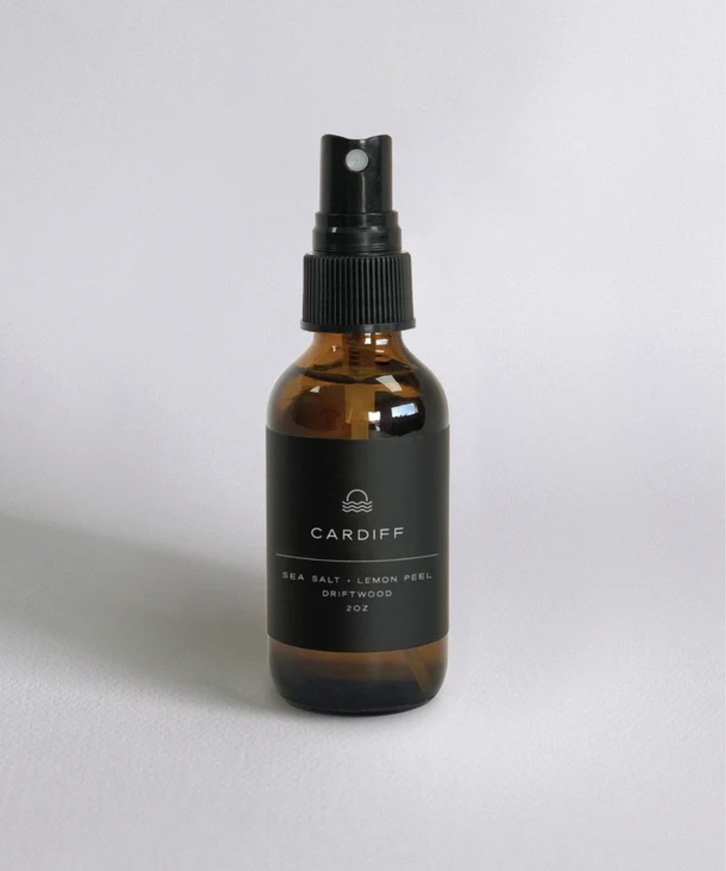 2oz amber spray bottle with black label reading "Cardiff: Sea Salt, Lemon Peel, Driftwood." Minimalist design, conveying a fresh, natural vibe.