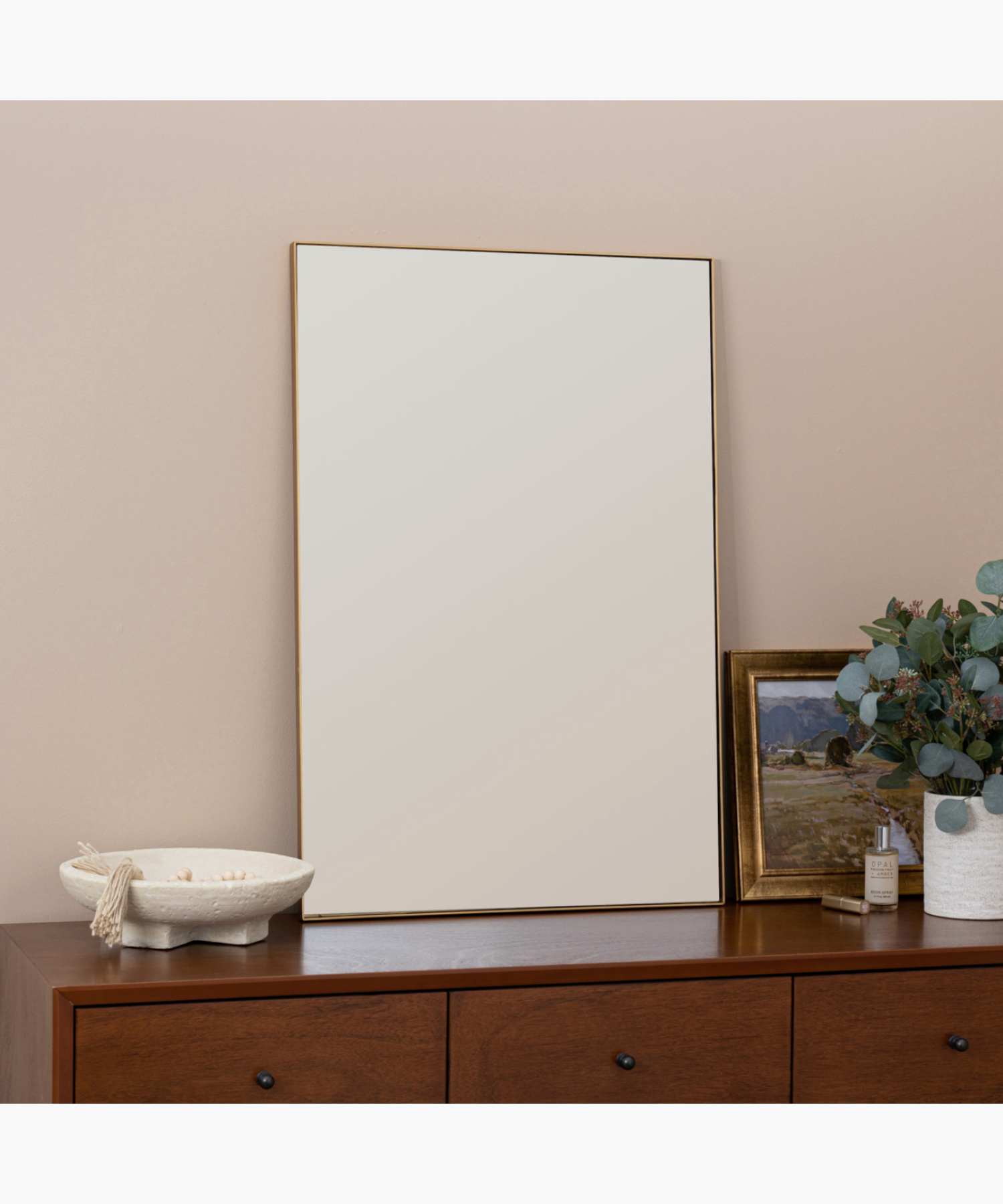 A minimalist living room features a large rectangular mirror on a wooden dresser. Beside it, a vase with eucalyptus, a landscape painting, and a candle.