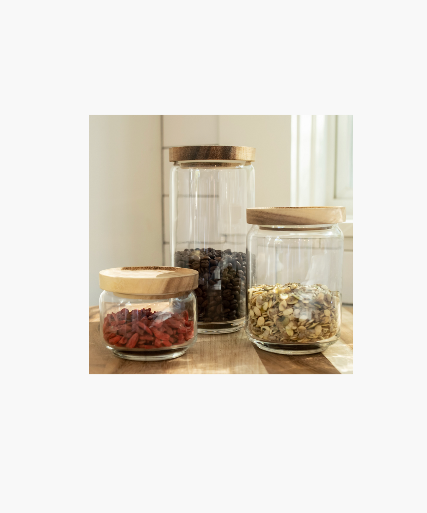 Three clear glass jars in various sizes each with a round, smooth wooden lid styled on a kitchen wooden kitchen counter. Each glass jar is filed with different dried food items. The simple, functional design conveys a minimalist aesthetic.