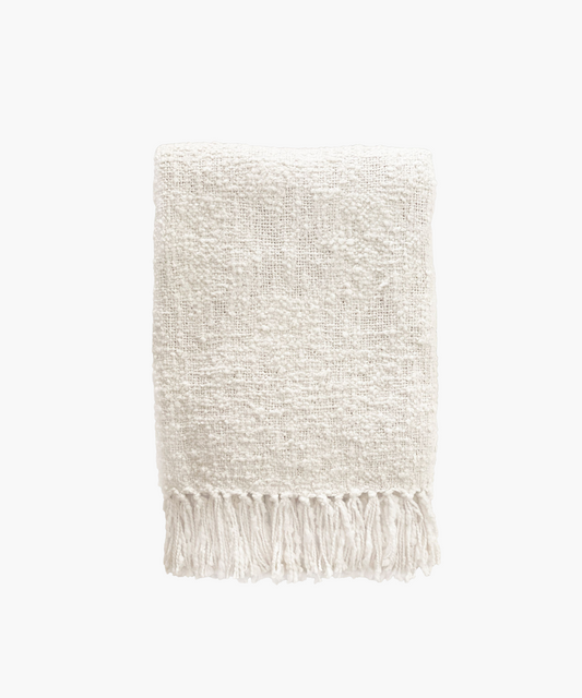 A soft, ivory colored woven throw blanket with a textured pattern and fringed edges, conveying a cozy and inviting feel. 