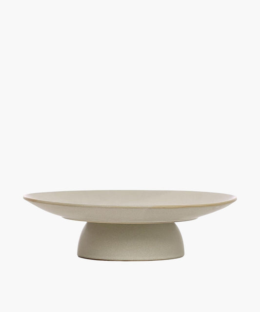 Minimalist, beige ceramic pedestal serving dish with a wide, flat surface on a sturdy base. It conveys simplicity and elegance in design.