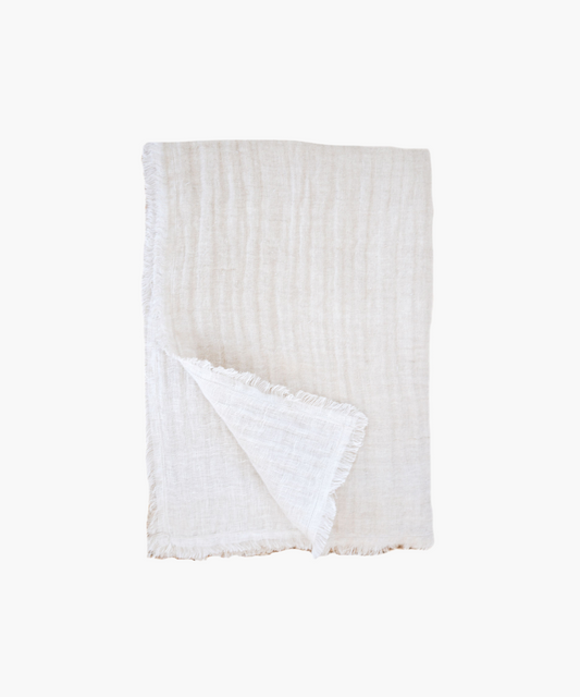 A soft, beige linen throw blanket is neatly folded against a plain background, showcasing a delicate fringe along the edges, conveying a cozy and elegant feel.