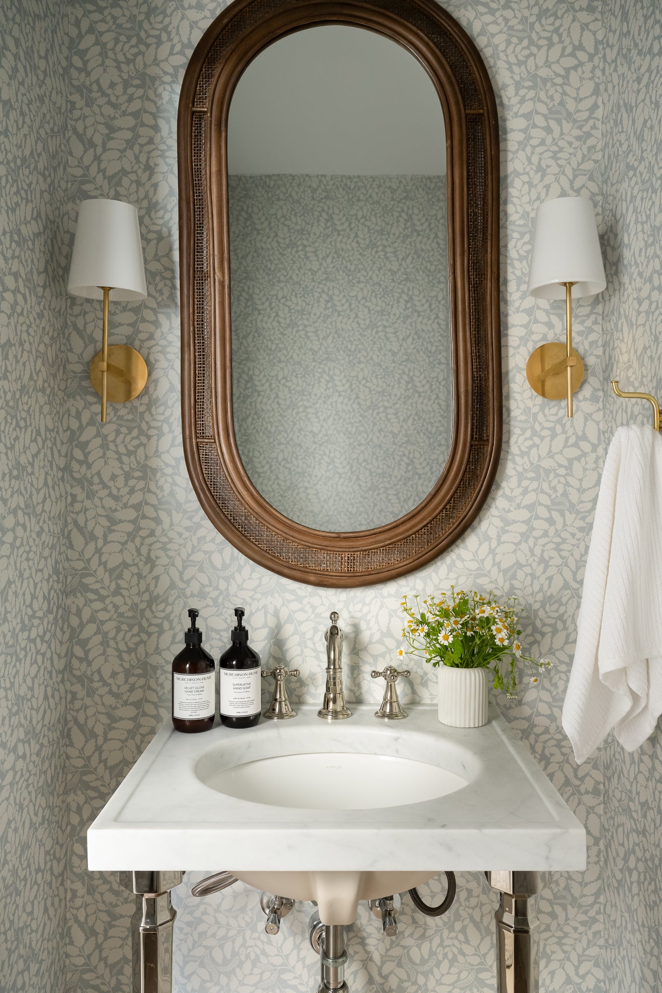 Elegant bathroom with a marble sink, decorative mirror, and floral wallpaper. Two sconces, hand soap bottles, and a vase with daisies add a refined touch.