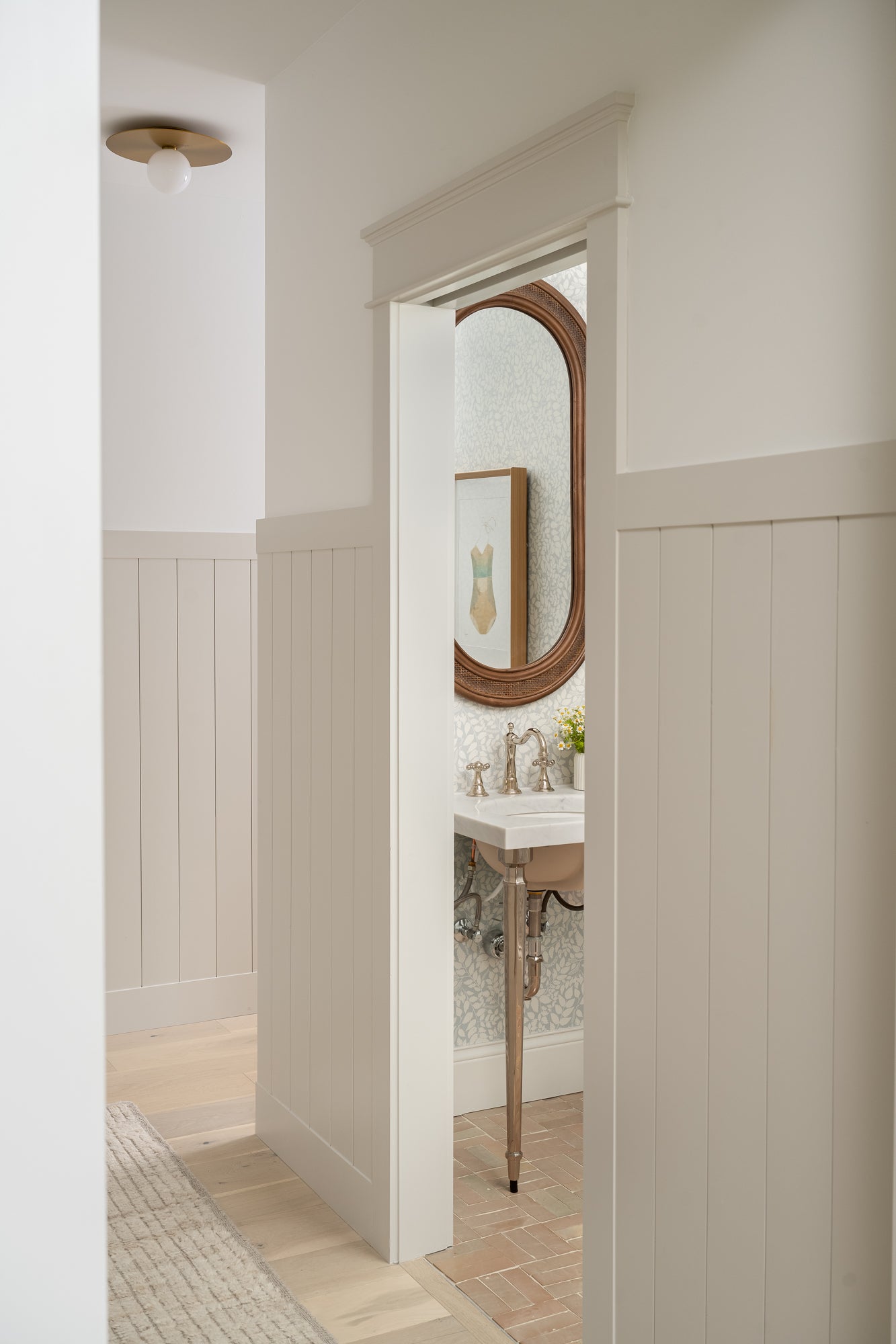 Hallway leading to a bathroom with white panel walls, a wooden floor, and a round mirror. The atmosphere feels clean and serene.