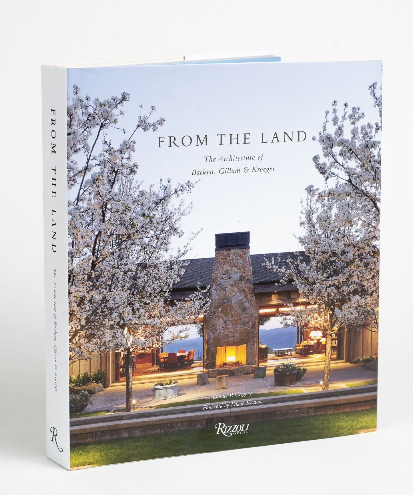 Cover of the book "From The Land" features an outdoor stone fireplace flanked by blossoming trees, with a warmly lit interior visible through large windows. Evening sky backdrop; peaceful ambiance.