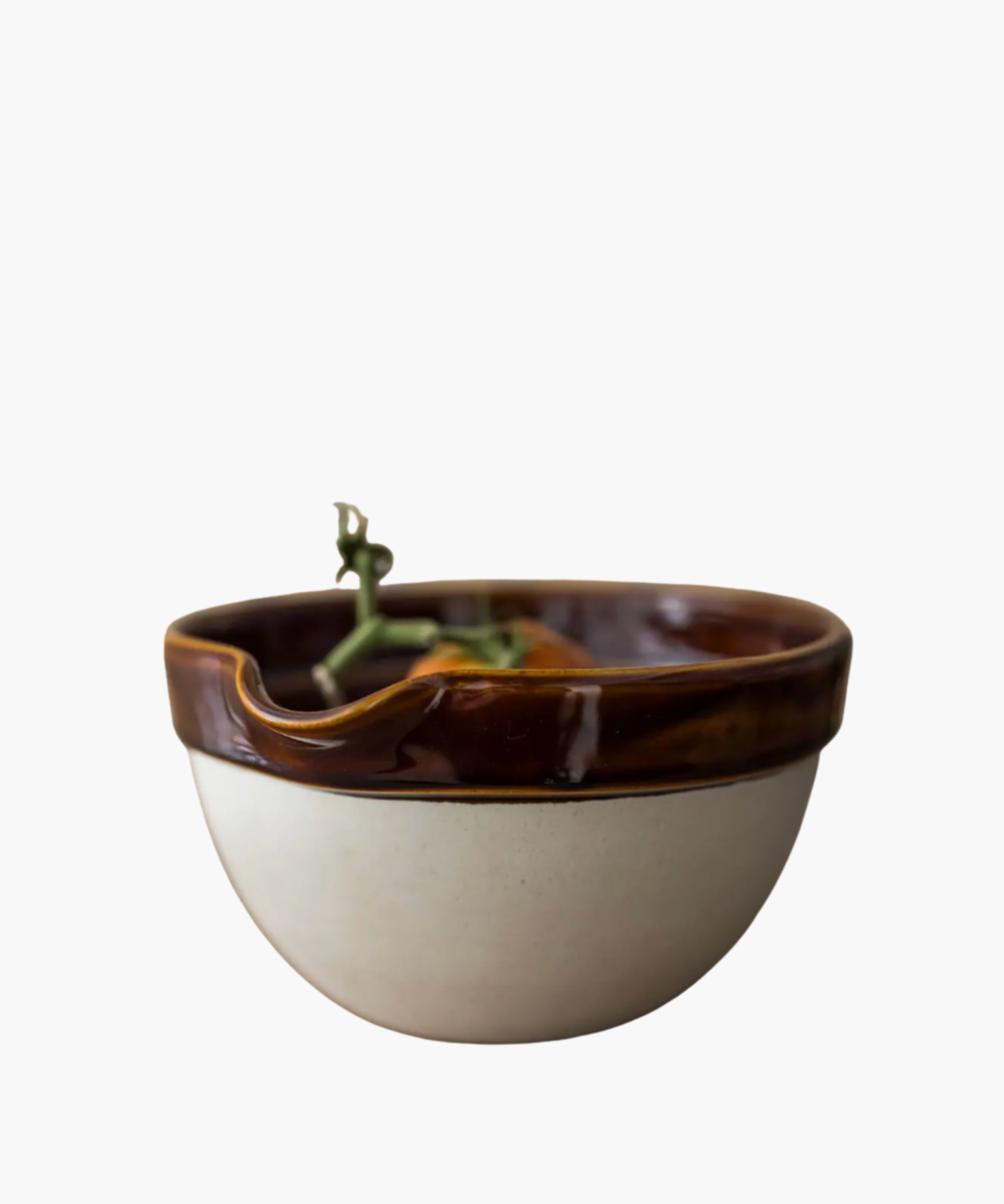 A ceramic bowl with a glossy brown rim and matte beige exterior holds a ripe tomato on a vine. The design is rustic and visually pleasing.