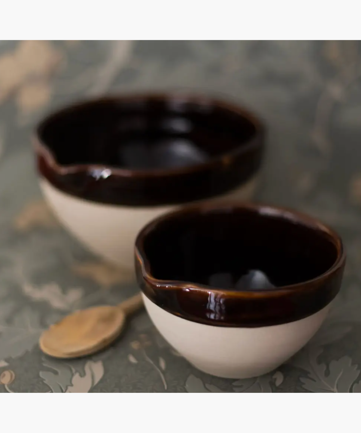 Two ceramic bowls with dark brown interiors and creamy exteriors sit on a floral patterned surface. A wooden spoon lies beside them, creating a rustic vibe.