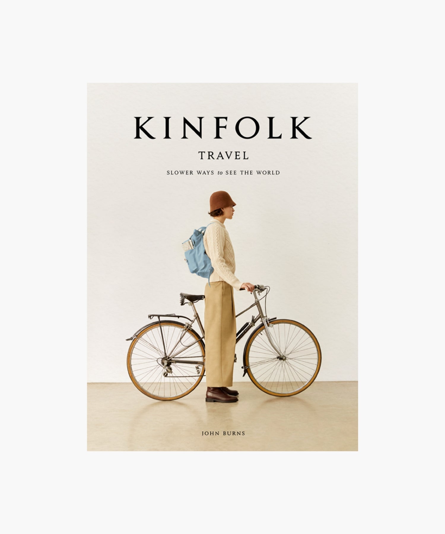 The cover of the book "Kinfolk Travel" by John Burns features a side view of a person in a brown hat, beige sweater, and khaki pants, standing next to a bicycle with a blue backpack. The tone is calm and nostalgic.