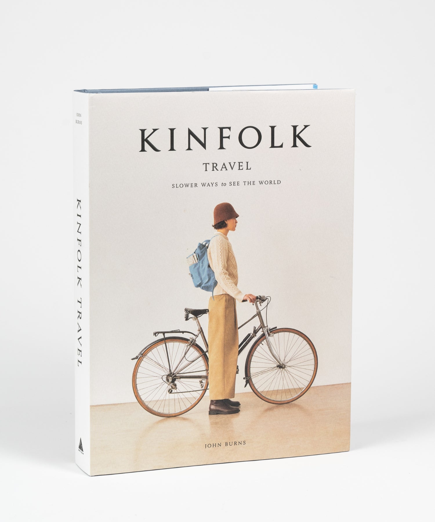 The cover of the book "Kinfolk Travel" by John Burns features a side view of a person in a brown hat, beige sweater, and khaki pants, standing next to a bicycle with a blue backpack. The tone is calm and nostalgic.