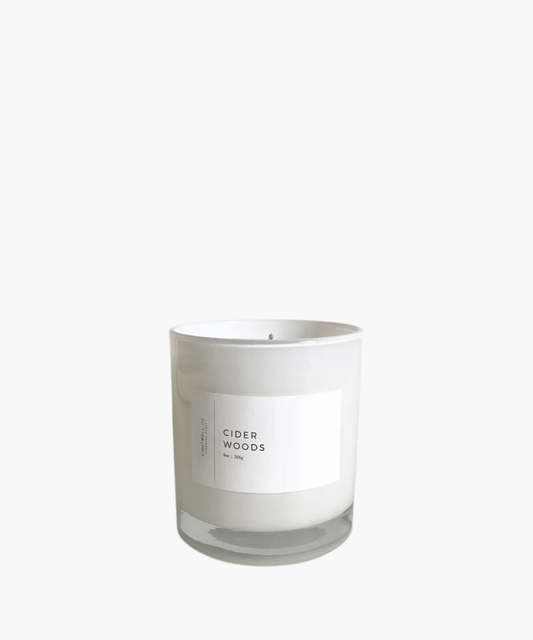 A white candle in a glass jar with a minimalist label reading "Cider Woods." The design conveys a clean, calming aesthetic.