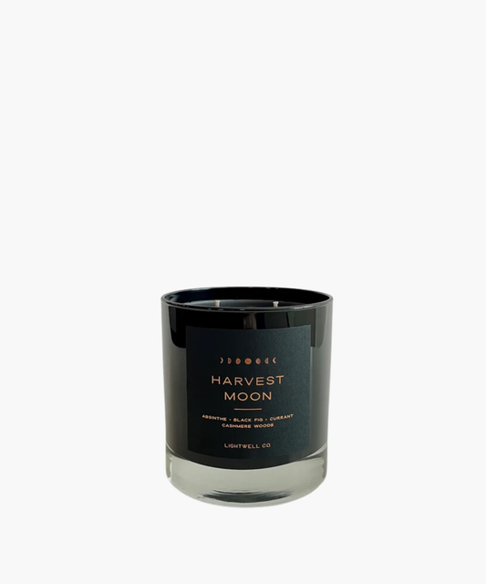 Black candle jar with a sleek design, labeled "Harvest Moon." Features a moon phase graphic and notes of absinthe, black fig, currant, and cashmere woods.