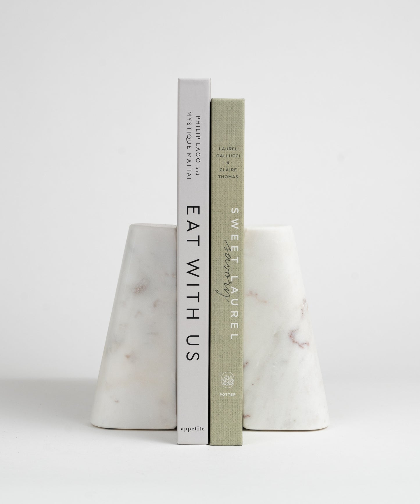 Two white marble bookends with a smooth, conical shape, each featuring subtle brown and gray veining styled with two cookbooks. They appear minimalist and elegant.