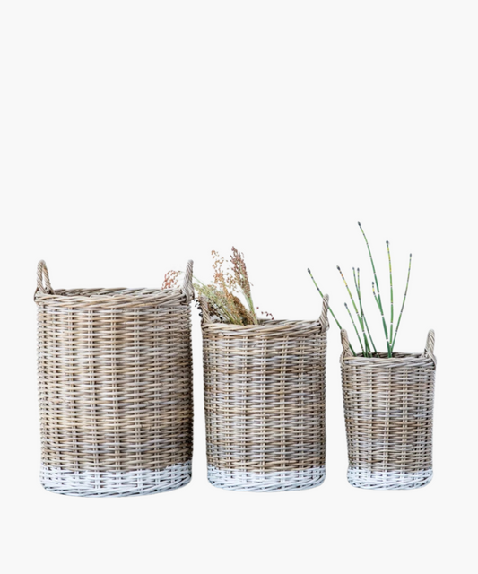 Rattan market baskets with a natural finish and a white dipped detail accenting the bottom in 3 sizes. Two baskets hold dried plants and sticks, conveying a rustic, natural vibe.