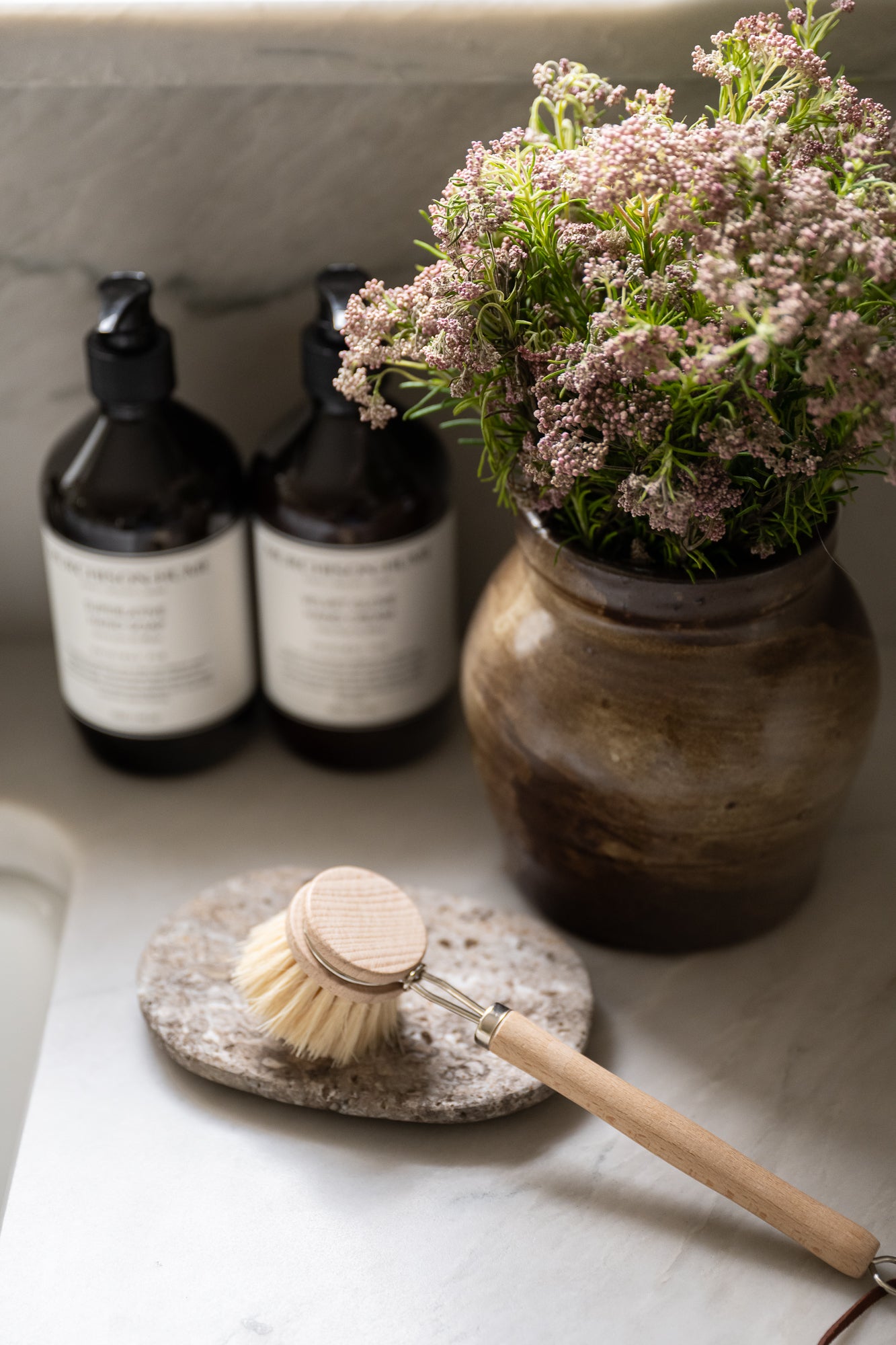 Oval shaped travertine soap dish with a rough, textured surface in shades of gray and hints of brown styled with kitchen wares. It conveys a rustic, natural, and minimalist aesthetic.