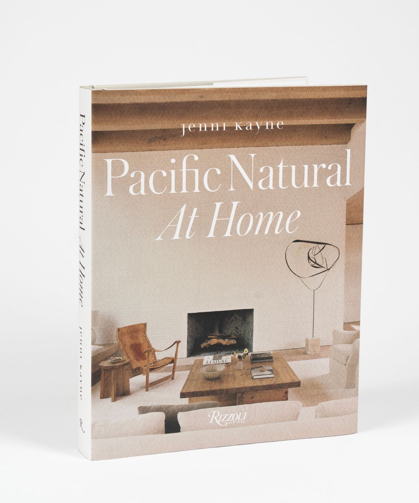 The cover of the book "Pacific Natural at Home" by Jenni Kayne features a living room with minimalist decor; features a white sofa, rustic wooden coffee table, leather chair, modern art piece, and a cozy fireplace. Serene, natural ambiance.