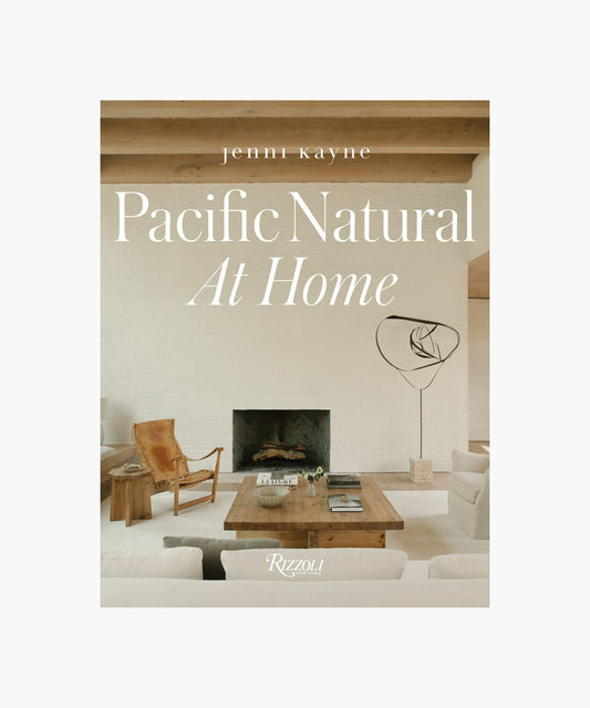 The cover of the book "Pacific Natural at Home" by Jenni Kayne features a living room with minimalist decor; features a white sofa, rustic wooden coffee table, leather chair, modern art piece, and a cozy fireplace. Serene, natural ambiance.