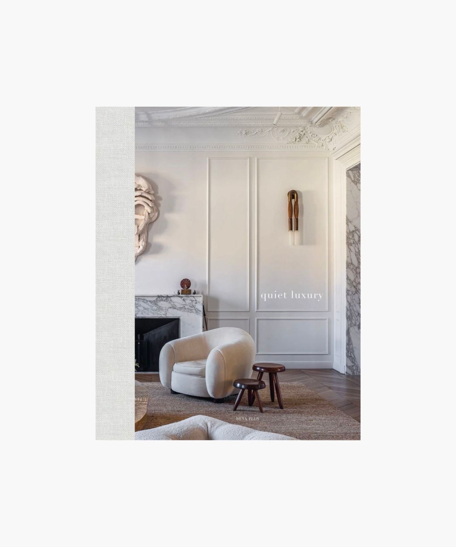 The cover of the book "Quiet Luxury" features a sophisticated living room with a plush beige armchair, two dark wood stools, ornate wall paneling, and a marble fireplace.