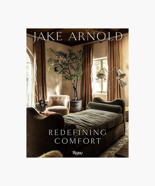 The cover of the book "Redefining Comfort" by Jake Arnold features a cozy living room with plush sofas, an elegant large potted plant, and soft lighting. Earthy tones create a warm, inviting ambiance with a touch of luxury.