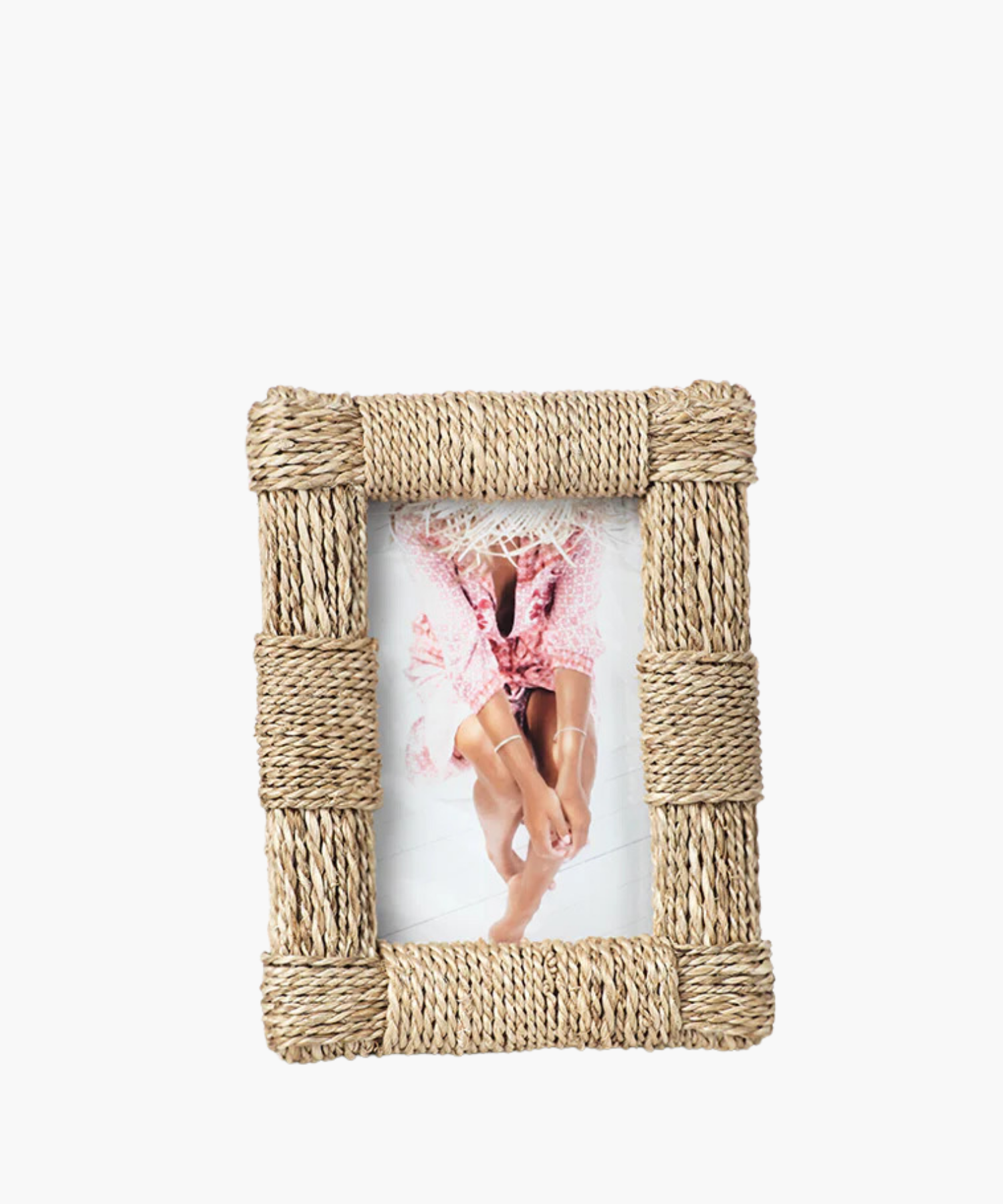 A rustic, woven rope frame with a picture of a person in a pink floral dress and straw hat sits on the floor, with legs bent and arms resting on knees. 