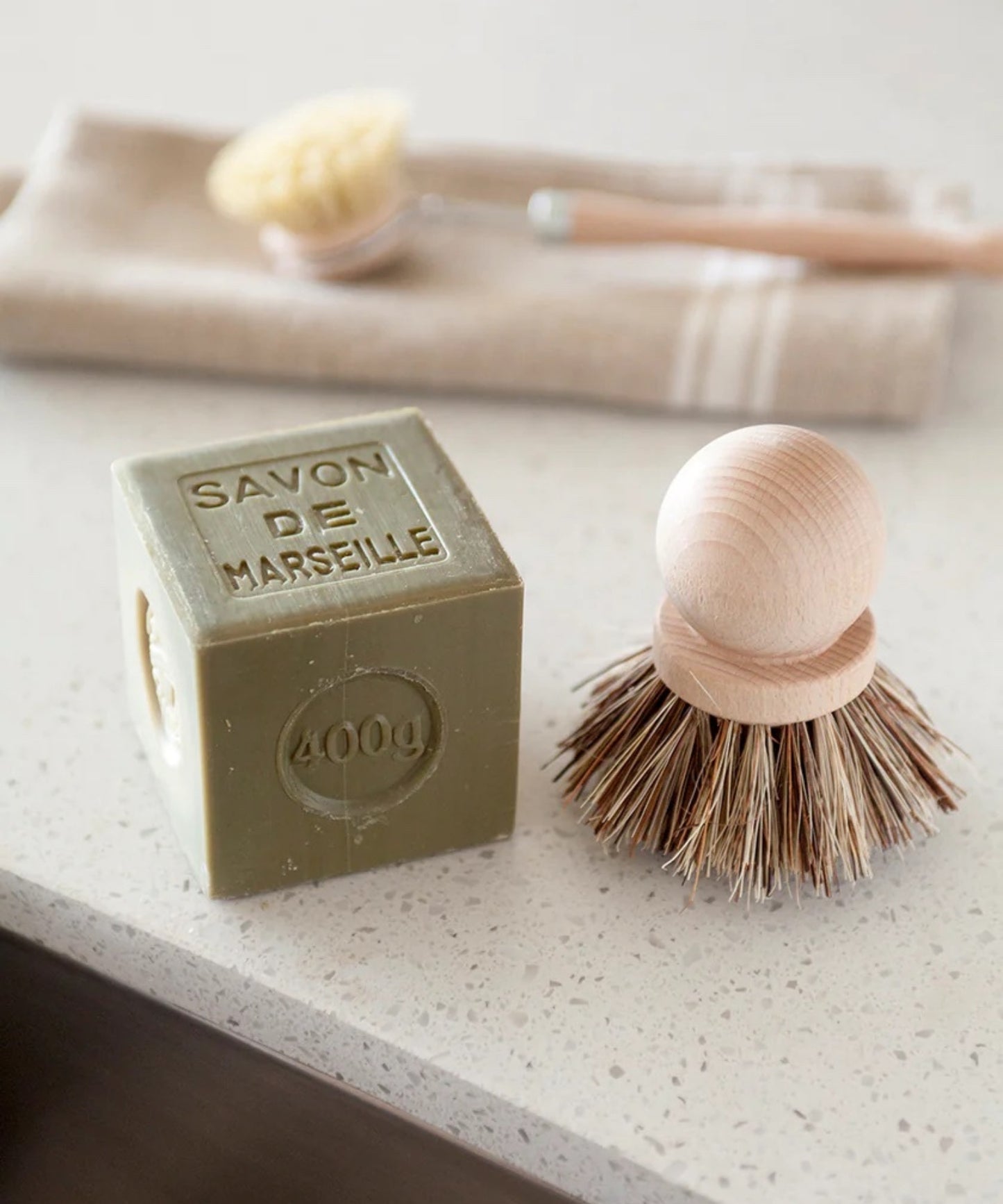 Wooden dish scrubber or saucepan brush with natural bristles, circular handle on top styled with a square block of soap. Simple, eco-friendly cleaning tool with a minimalistic vibe.