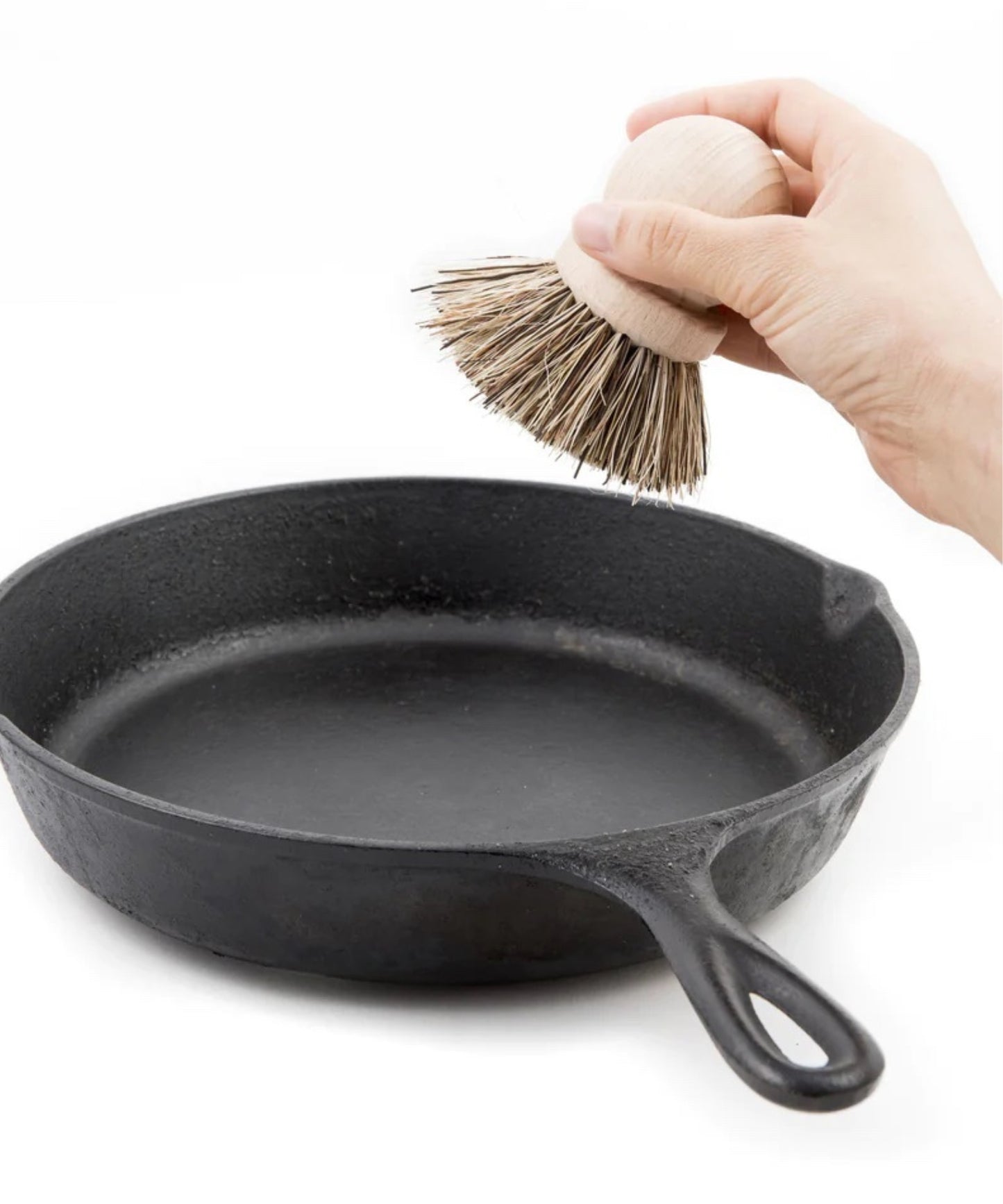 Wooden dish scrubber or saucepan brush with natural bristles, circular handle on top styled with a cast iron skillet. Simple, eco-friendly cleaning tool with a minimalistic vibe.