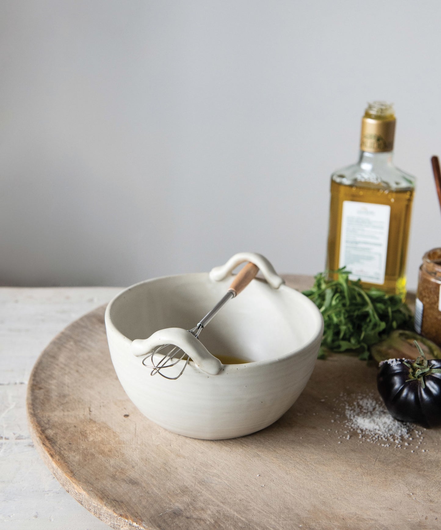 A cream colored stoneware ceramic mixing bowl with a metal whisk styled with kitchen ware. Evoking a clean, minimalist aesthetic.