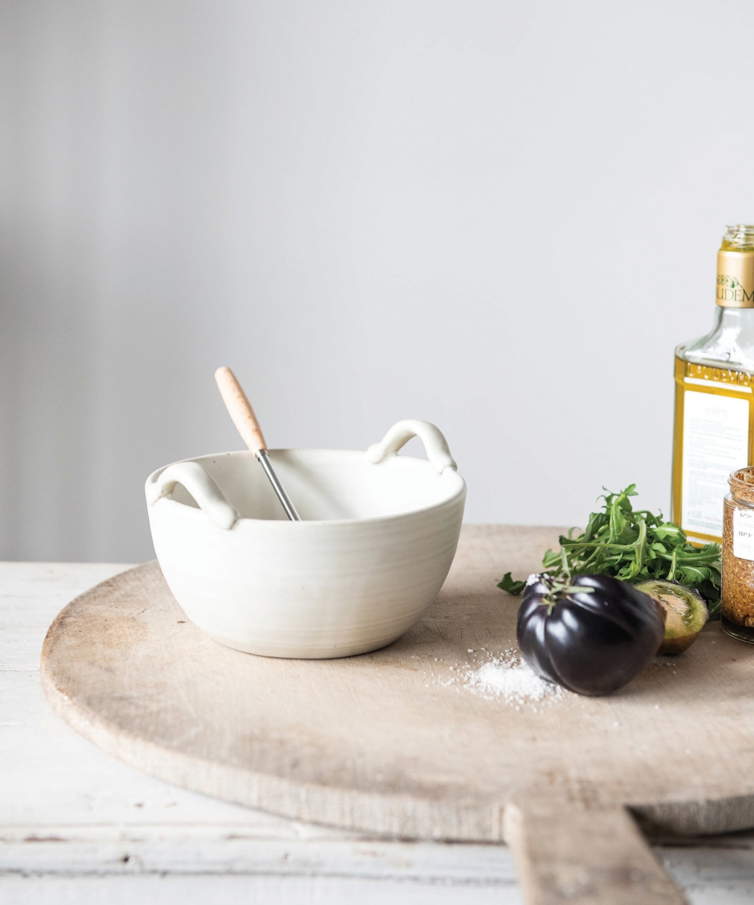 A cream colored stoneware ceramic mixing bowl with a metal whisk styled with kitchen ware. Evoking a clean, minimalist aesthetic.