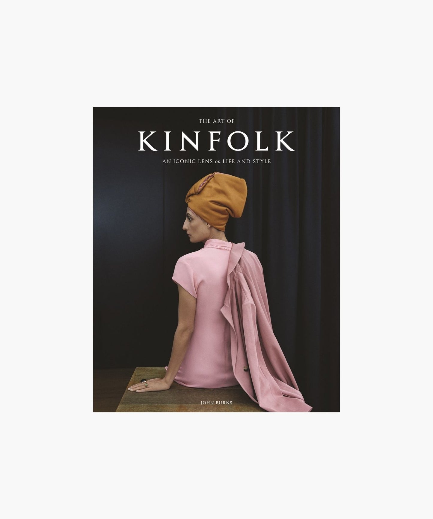 The cover of the book "The Art of Kinfolk" by John Burns features a woman in a pink dress and orange headwrap, sitting elegantly against a dark curtain. The tone is stylish and serene.