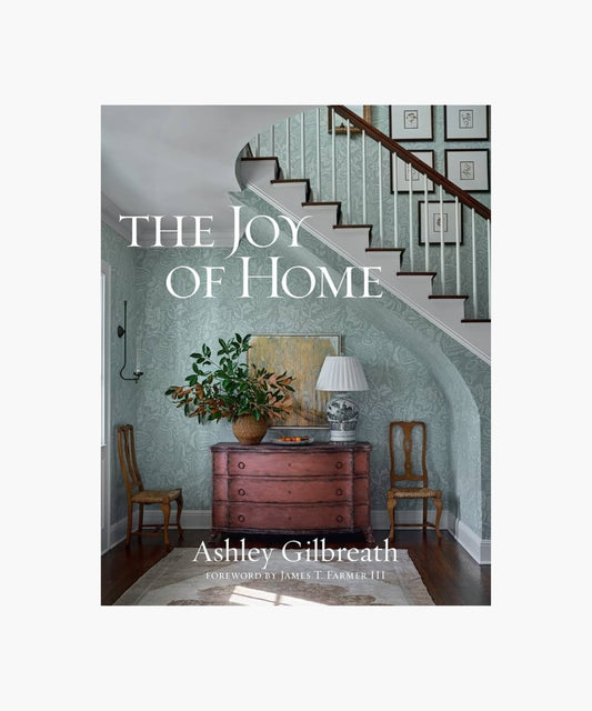 The cover of the book "The Joy of Home" by Ashley Gilbreath features a cozy, elegant hallway with a wooden chest, potted plant, and lamp beneath a staircase; framed prints on the staircase wall. Classic and inviting.