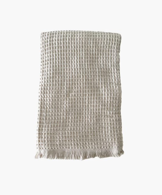 Beige waffle-textured throw blanket with fringed edges, laid flat. The soft material conveys a cozy, inviting feel. Suitable for any living room or bedroom.