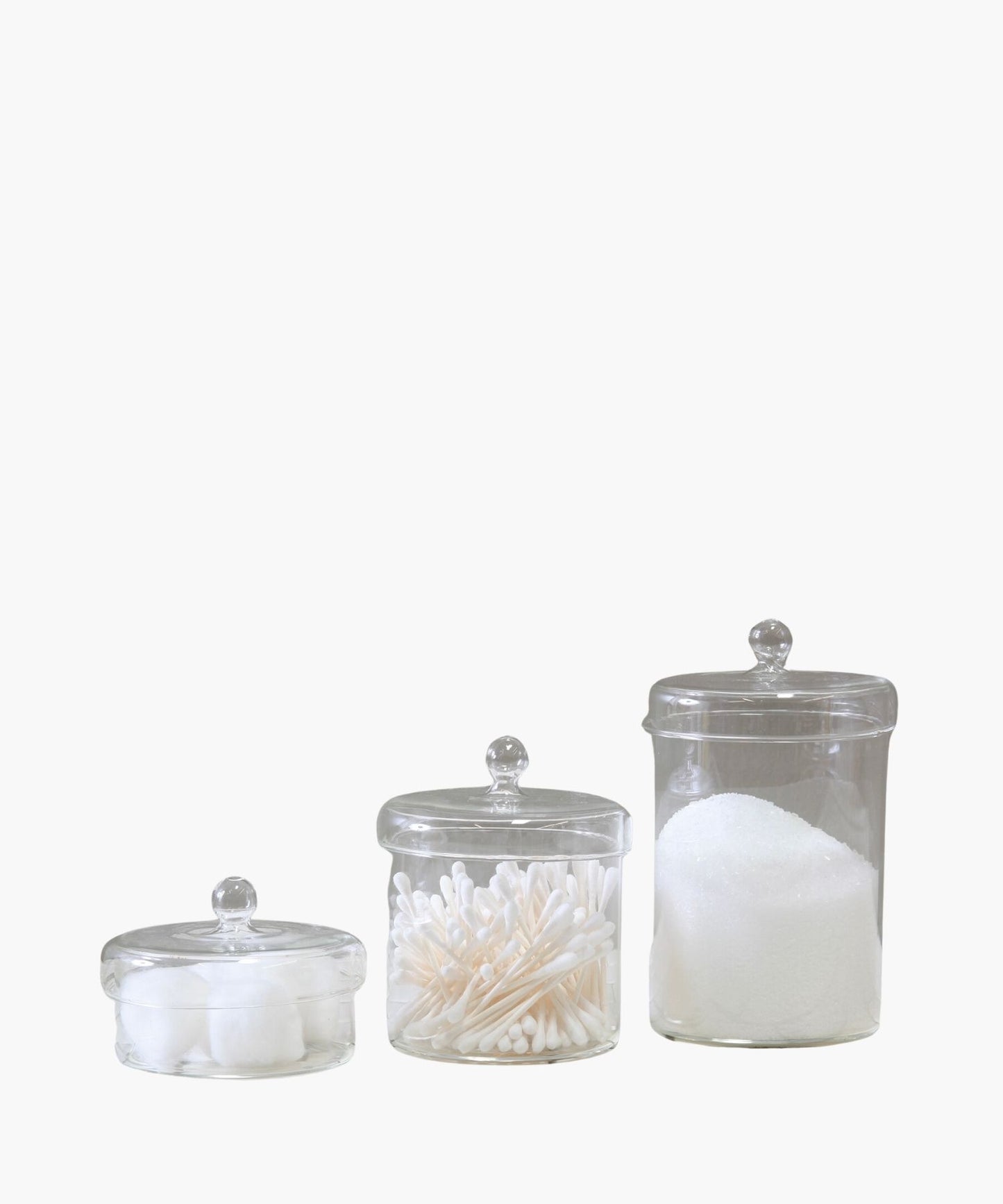 Three clear glass jars in different sizes with each with a round lid and a knob handle is centered on a plain white background. The jars are filled with various bathroom accessories.