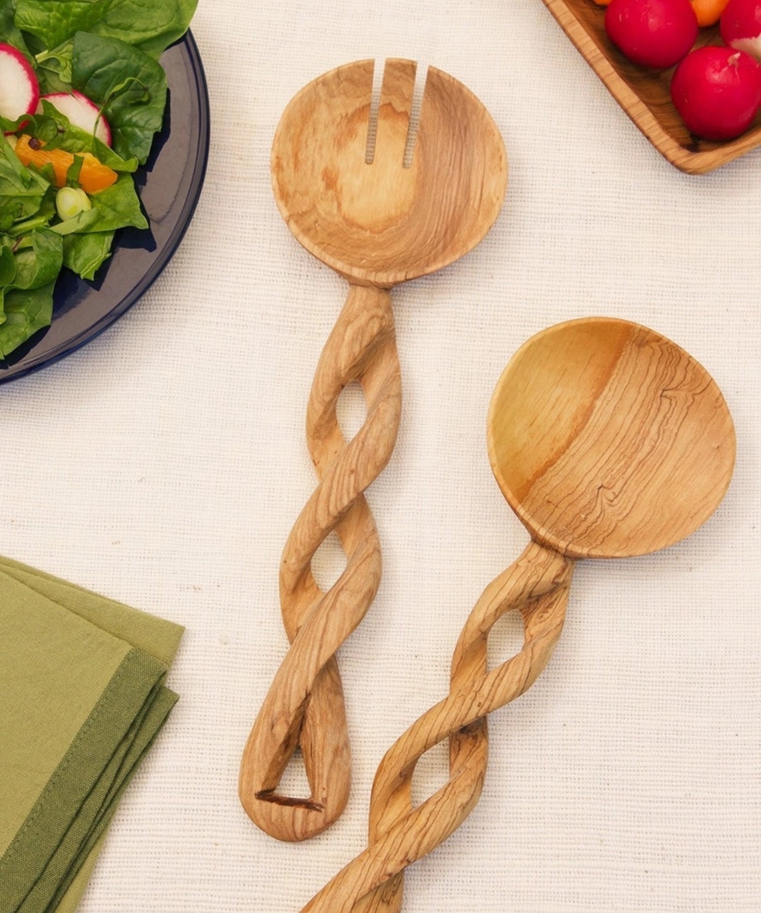 Two ornately carved olive wood salad servers, a spoon and a fork, featuring twisting handles and rich natural wood patterns. They convey a rustic, artistic feel.