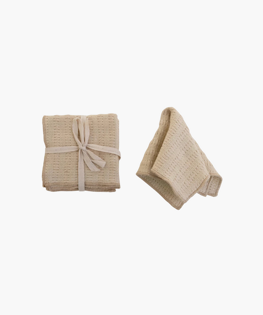 Two ivory waffle-textured dish cloths; one folded and tied with a white ribbon, the other loosely folded. The tone is neutral and cozy.