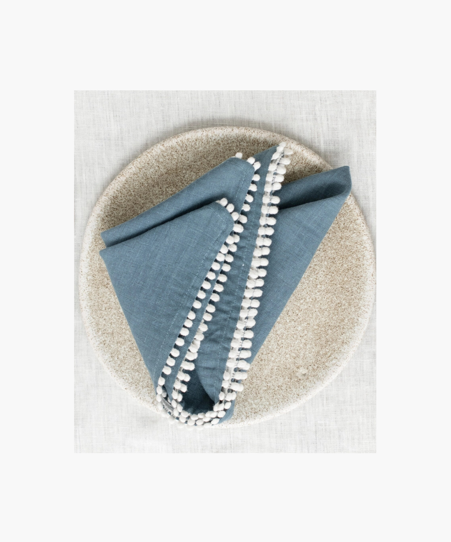 A blue linen napkin with white pom-pom trim neatly folded on a speckled ceramic plate, set against a light, textured fabric background. The scene is elegant and minimalistic.