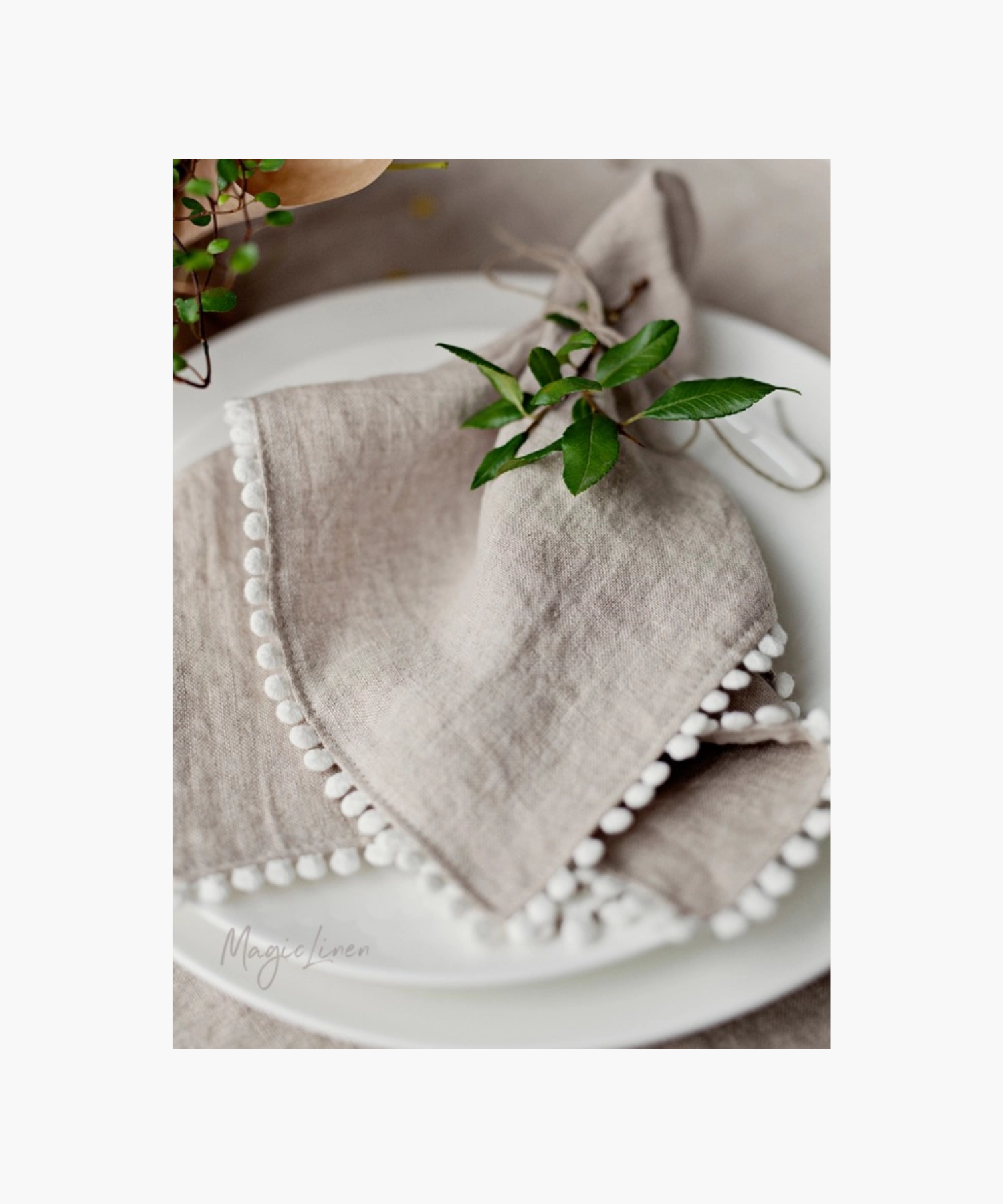 Elegant table setting featuring a delicate beige linen napkin with white pom-pom trim, accented by fresh green leaves on a white plate.