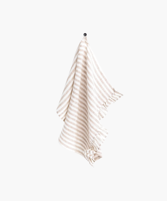 A beige and white striped linen tea towel with ruffle trim hangs neatly on a black hook against a plain white background, creating a clean and minimalistic appearance.